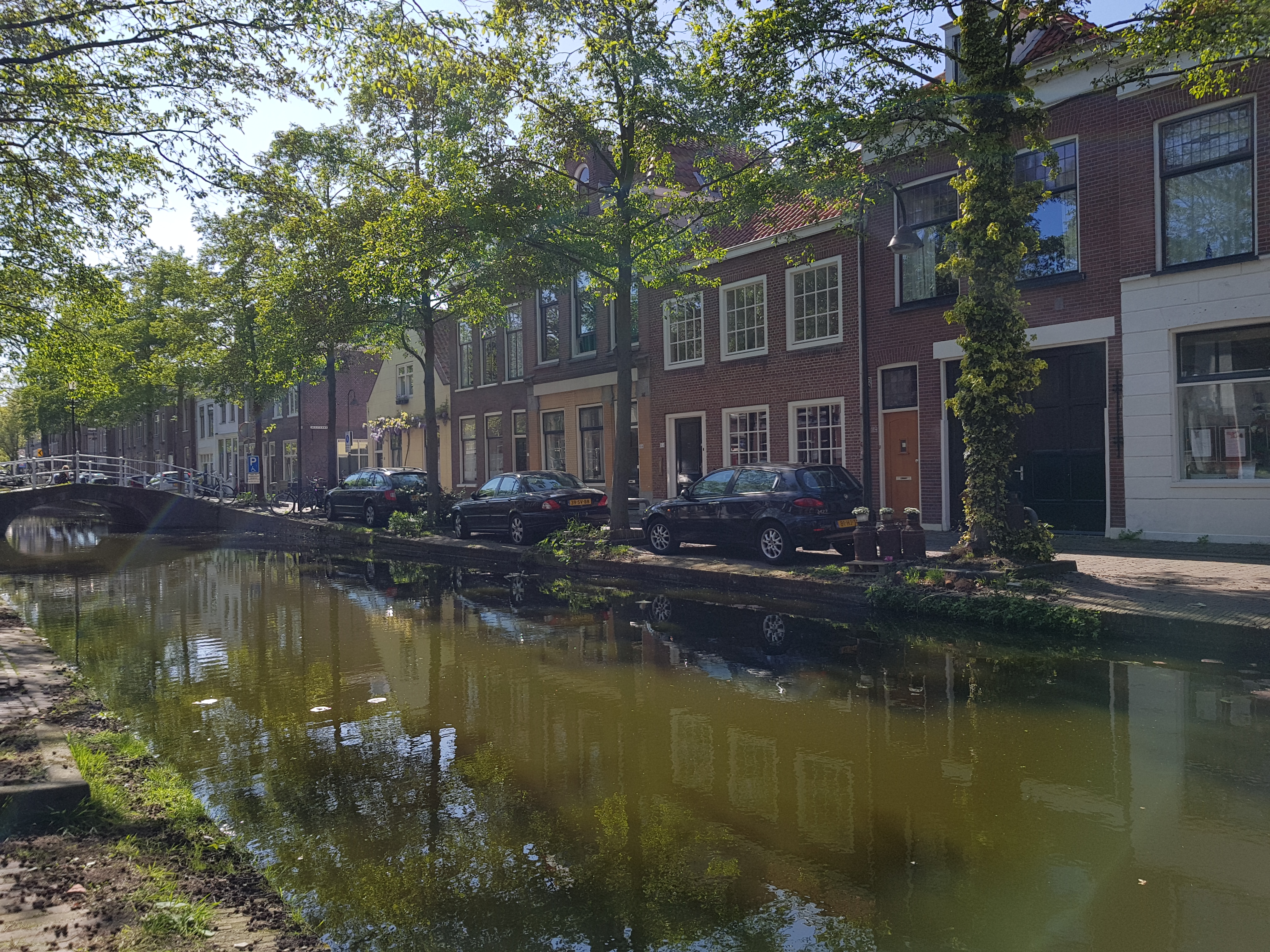 Dutch-style historic row houses along a canal with green water and blue skies with white clouds. Cars are parked along the canal.